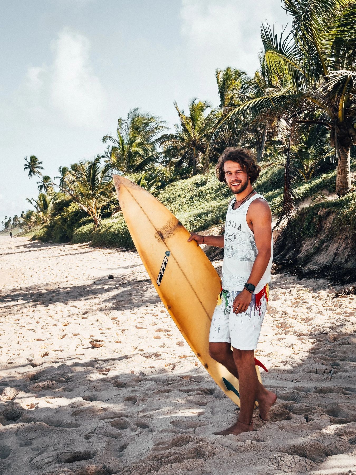 Lucas Kampf mit Surfboard an einem Palmenstrand
